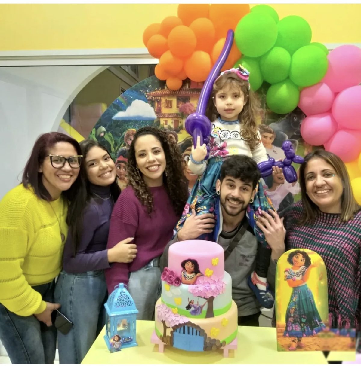 Equipe com a aniversariante e o bolo, entre arcos de balões